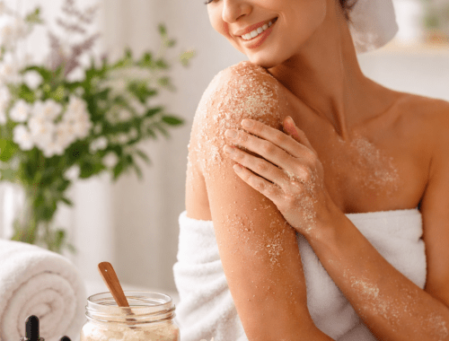 Natural skincare routine with a woman using an essential oil body scrub surrounded by essential oil bottles and fresh flowers.